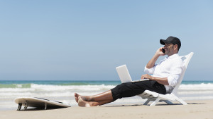 Um homem com celular e computador trabalhando na praia